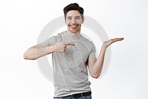 Image of handsome smiling man pointing at his empty hand, holding item on palm, display your product, demonstrate copy