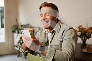 Image of handsome young man in cafe, looking at his mobile phone, using smartphone app, drinking coffee
