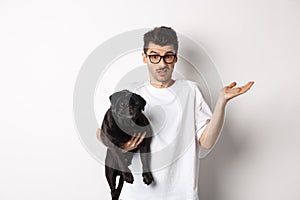 Image of handsome young man holding black pug and looking confused. Guy shrugging shoulders and staring indecisive at