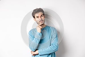 Image of handsome young man making choice, thinking and looking pensive up, standing over white background