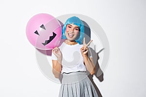 Image of happy asian girl celebrating halloween in blue anime wig, holding pink balloon with scary face and showing