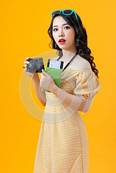 Image of happy asian young woman tourist standing isolated over yellow background holding camera and passport with tickets