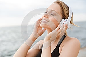 Image of happy beautiful woman listening to music with headphones near seaside in morning