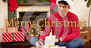 Image of happy holidays text over caucasian man writing christmas cards with presents