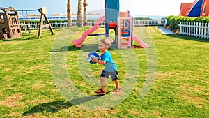 Image of happy laughing cheerful boy holding football ball in hands and running on children playground at park