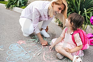 Image of happy little girl and mother laughing and drawing with colorful chalks on the sidewalk. Caucasian female play together