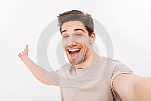 Image of happy man in casual t-shirt and bristle on face rejoicing and taking selfie with smile, isolated over white background