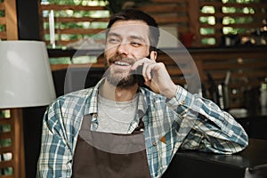 Image of happy waiter boy sitting at the bar and using cellphone while working in cafe or coffeehouse outdoor
