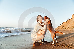 Image of happy woman 20s hugging her dog, while walking along the beach