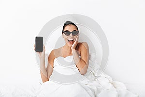 Image of happy woman 30s wearing sunglasses wrapped in blanket lying in bed and holding mobile phone