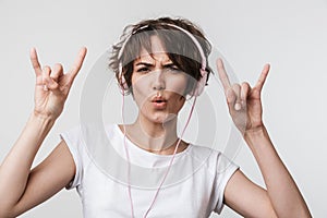 Image of happy woman in basic t-shirt showing rock sign while listening to music with headphones