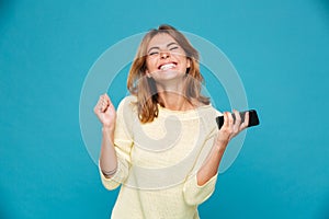 Image of Happy woman in sweater holding smartphone and rejoice