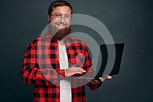 Image of happy young bearded man standing over grey wall background isolated. Looking camera using laptop
