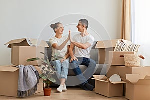 Image of happy young family buying new flat and posing surrounded with boxes with personal pile, sitting on sofa and giving five