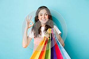 Image of happy young glamour woman shop in stores with discounts, showing okay sign, holding shopping bags, looking