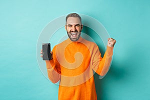 Image of happy young man winning and showing mobile phone screen, rejoicing and celebrating victory, fist pump with