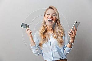 Happy young pretty blonde business woman posing isolated grey wall background holding credit card using mobile phone photo
