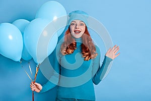 Image of happy young woman celebrating birthday with air balloons, looking at camera and raising hand, wearing casual shirt and