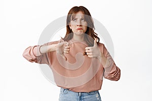 Image of hesitant young woman thinking while showing thumbs up down, pondering while leaving review feedback, standing