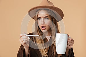 Image of ill caucasian girl holding thermometer and hot tea cup