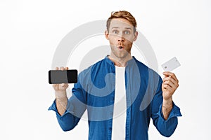 Image of impressed young man shows horizontal mobile phone screen and credit card, gasping and saying wow, new bank