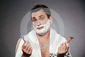 Image of indecisive man with shaving foam on his face holding two razors, isolated over gray background