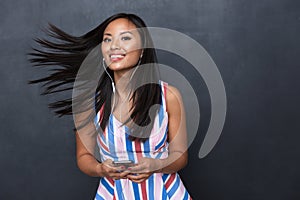 Image of joyful asian woman 30s wearing earphones listening to music on black smartphone standing isolated