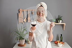 Image of joyful attractive winsome woman with cosmetic mask in white towel wrapped around head drinking wine in kitchen, rejoicing