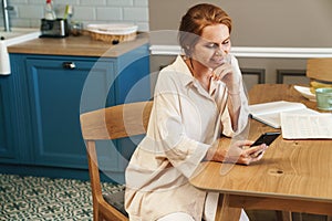 Image of joyful ginger woman using cellphone while siting at table