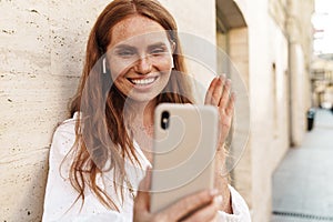 Image of joyful ginger woman waving hand and using cellphone