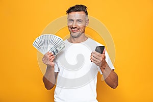 Image of joyful guy 30s in white t-shirt holding cell phone and bunch of money