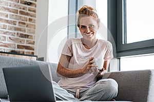 Image of joyful redhead woman drinking coffee and using laptop