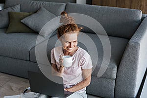 Image of joyful redhead woman drinking coffee and using laptop