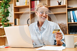 Image of joyful woman holding credit card while working with laptop