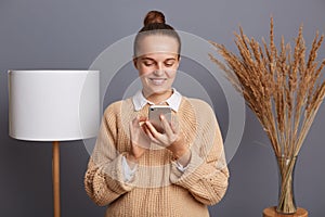 Image of joyful woman wearing warm sweater using cell phone for checking cocial networks, expressing positive emotions, standing