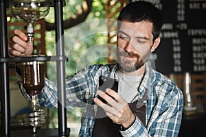 Image of joyous barista boy using cellphone and making coffee while working in cafe or coffeehouse outdoor