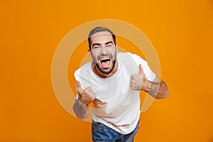 Image of joyous guy 30s in t-shirt rejoicing and showing thumbs up while standing,  over yellow background