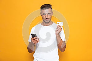 Image of joyous guy 30s in white t-shirt holding mobile phone and credit card