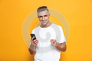 Image of joyous man 30s in white t-shirt smiling and holding smartphone while standing isolated