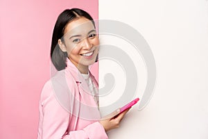 Image of korean female entrepreneur in suit, standing near info wall, advertisement on board, holding smartphone and