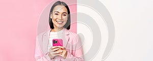 Image of korean female entrepreneur in suit, standing near info wall, advertisement on board, holding smartphone and