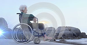 Image of light moving over smiling senior caucasian woman in wheelchair on beach looking to sea