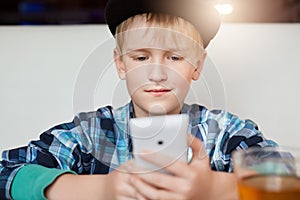 Image of little blonde boy with blue eyes dressed in shirt in a cage print and stylish cap siting at cosy restaurant using mobile