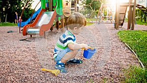 Image of little boy sitting on the playground and puring sand with small plastic spade in colorful bucket. Kid digging
