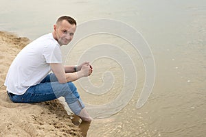 The image lonely positive and smiling man sitting on a beach by the river.