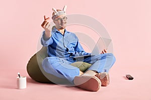 Image of man sitting on bag chair in pajama and funny headband with laptop with love-sign over pastel rose background. photo