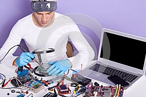 Image of man using magnifier while looking on wires of pc, concentrated male working in electronic lab, sits at table surrounded