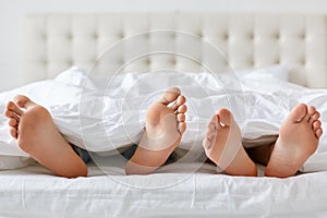 Image of man and woman bare feet under blanket in bedroom. Unrecognizable husband and wife spend free time in comfortable bed, enj