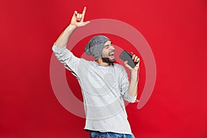 Image of optimistic man 30s singing while listening to music with earphones and mobile phone, isolated over red background