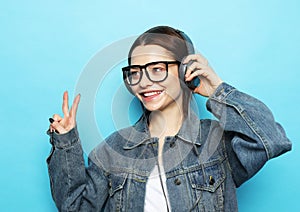 Image of optimistic woman wearing glasses listening to music with headphones and show victory sign